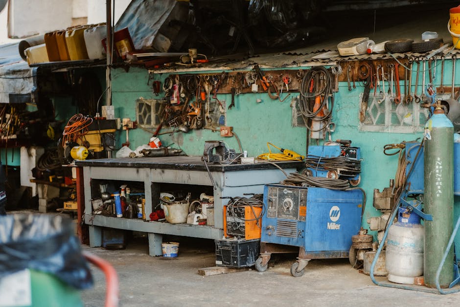 A cluttered workbench situated in a garage workshop with a turquoise painted wall behind it. The workbench surface is made of wood and is covered with various tools, including pliers, screwdrivers, an