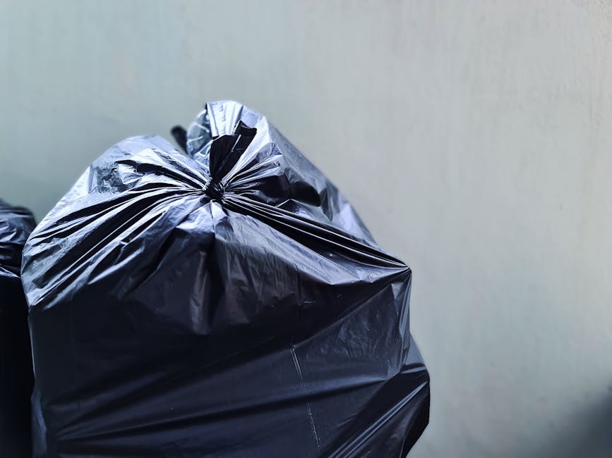 A close-up view of two black rubbish bags made of crinkled, shiny plastic material, stacked on top of each other against a plain, light-colored wall background. The bags are tied at the top with visib