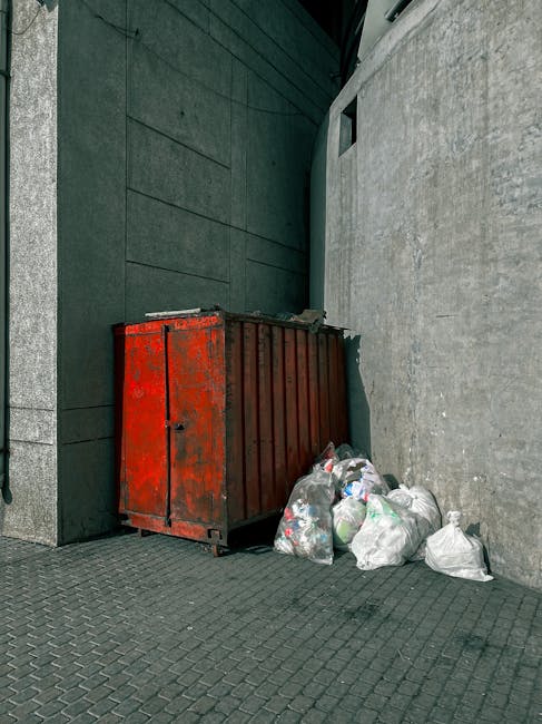 A close-up view of two black rubbish bags made of crinkled, shiny plastic material, stacked on top of each other against a plain, light-colored wall background. The bags are tied at the top with visib