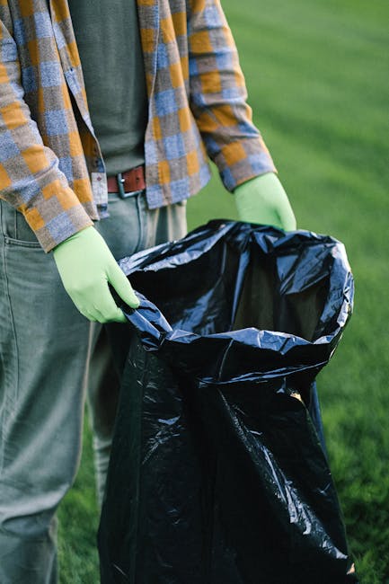 Two sanitation workers wearing dark uniforms with reflective orange and yellow stripes are collecting waste in an urban area during the evening, standing beside a large white rubbish collection truck.
