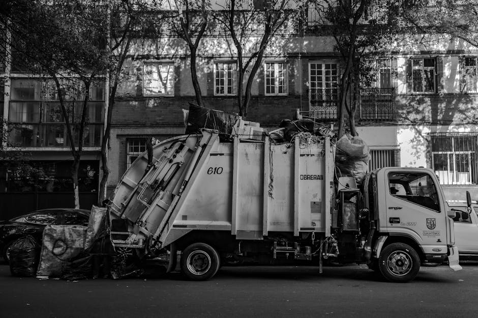 A sanitation worker dressed in a high-visibility uniform with reflective stripes is seen walking across a paved town square, pushing a large black wheeled rubbish bin on its side. The worker is wearin
