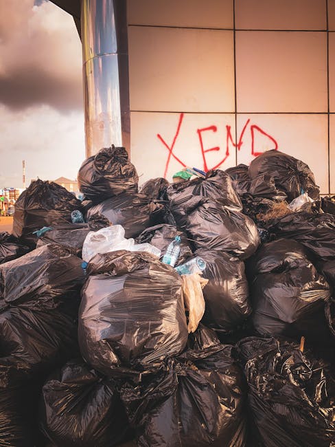 A large pile of black rubbish bags, filled and tied, is stacked against a tiled wall outdoors. Some of the bags are slightly torn or bulging, revealing mixed waste inside. Among the bags, several clea
